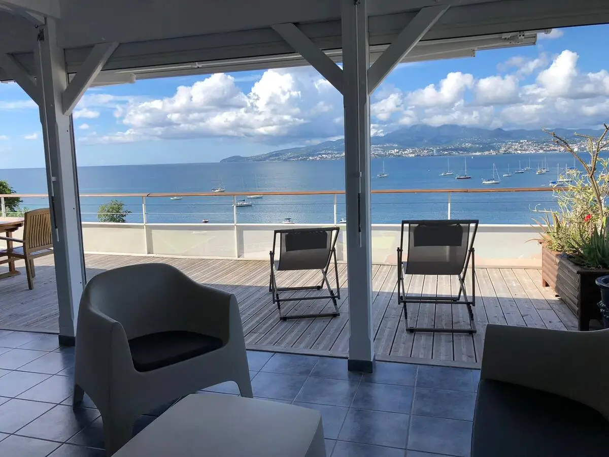 Vue depuis le salon ouvert sur la terrasse panoramique avec la baie de Fort-de-France en arrière-plan - Appartement Grand Large Trois Îlets Martinique