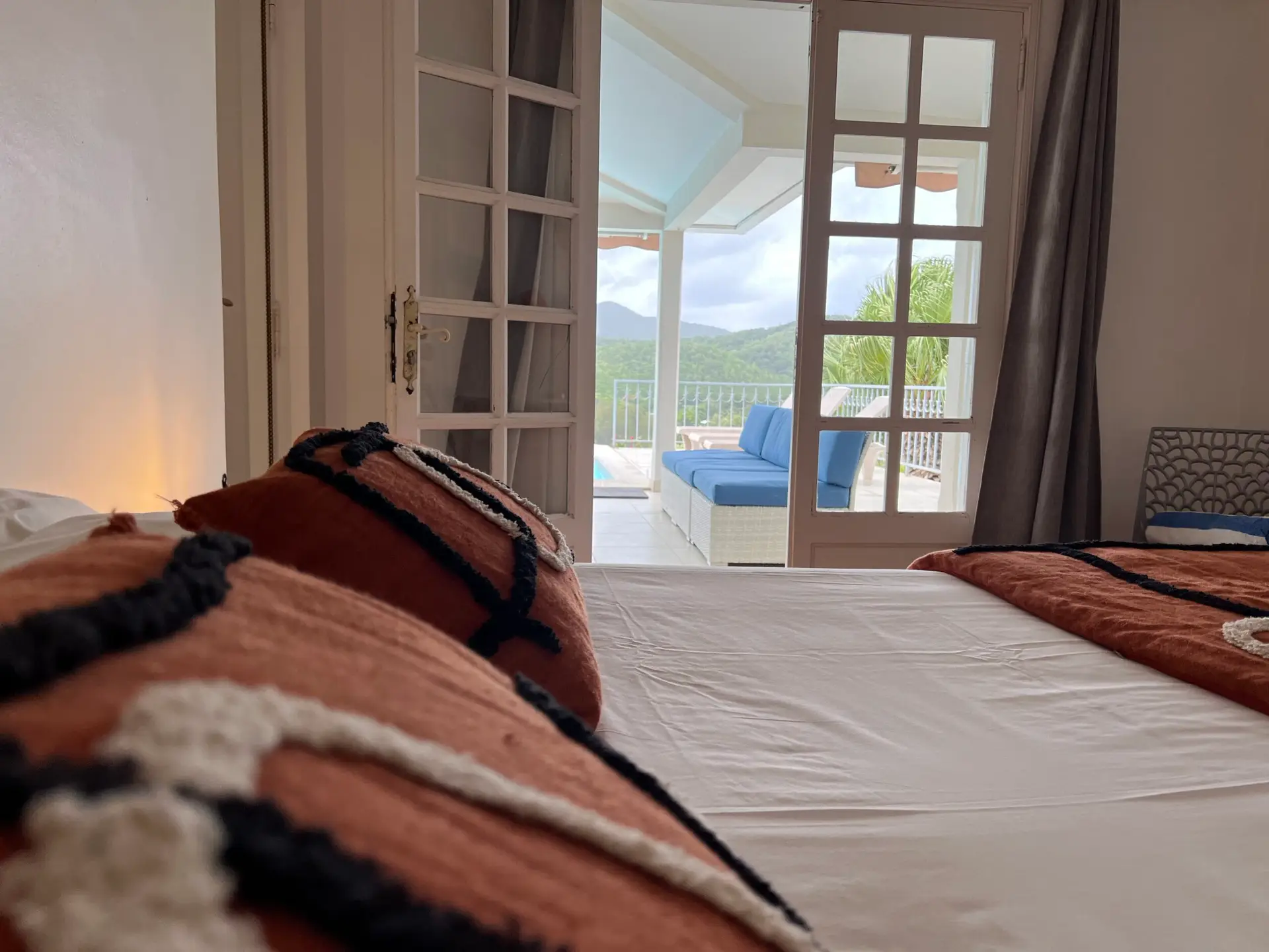 Vue depuis le lit d’une chambre de la Villa Thalizia en Martinique, donnant sur la terrasse et la piscine avec panorama sur les collines.